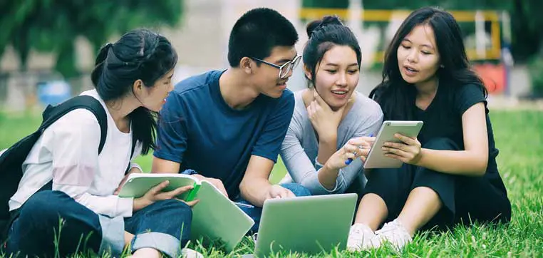 Four young adults, possibly exploring colleges for Asian students, sit on the grass, engaging with laptops and a tablet in an outdoor setting.