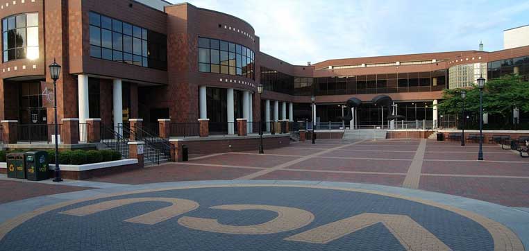 A brick courtyard with the letters "VCU" on the ground is surrounded by a large brick building with multiple windows and white columns, embodying the spirit of Virginia Commonwealth University.