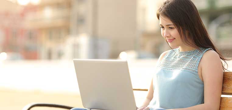 A woman in a sleeveless top is sitting on a bench and using a laptop outdoors, exploring college visit alternatives.