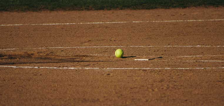 A yellow softball sits on the dirt infield near home plate on a softball field, typical of the facilities found at many NCAA D2 colleges.