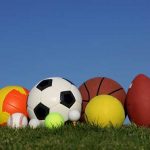 A collection of sports balls, including a soccer ball, basketball, and football, arranged on grass against a clear blue sky, often used in athletic recruiting.