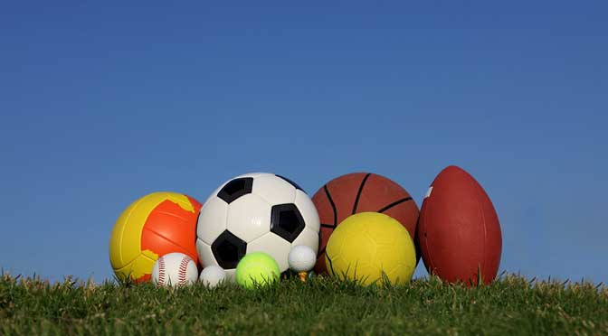 A collection of sports balls, including a soccer ball, basketball, and football, arranged on grass against a clear blue sky, often used in athletic recruiting.