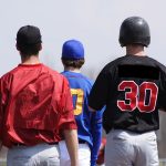 Three baseball players, seen from behind, walk together. These athletes, important to college baseball coaches, wear different jerseys: the left in red, the middle in blue, and the right in black with the number 30 on the back.