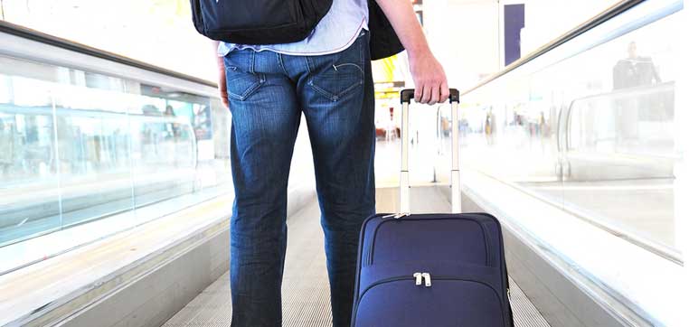 A person wearing jeans and a white shirt walks on an airport travelator while pulling a navy blue suitcase and carrying a black backpack, perhaps heading to one of the colleges with the most geographic diversity.