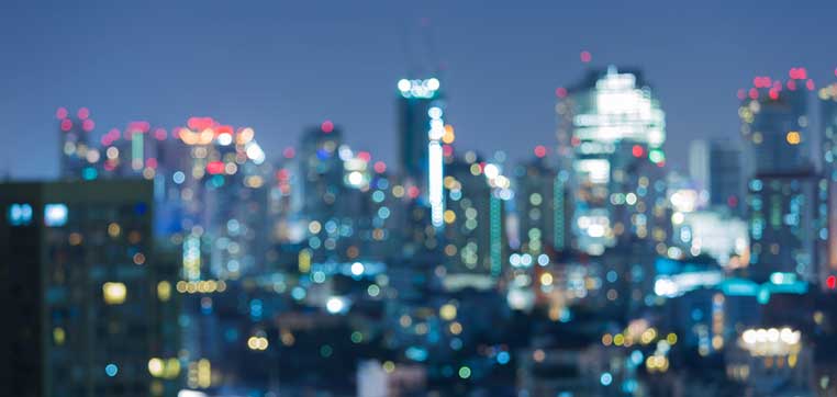 Blurry image of a city skyline at night with numerous lights and illuminated buildings against a dark sky, reminiscent of bustling colleges in big cities.