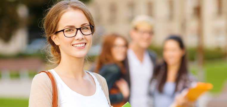 A young woman in glasses and a white top smiles at the camera, holding a green folder that contains a list of flagship universities. Several people stand blurred in the background.