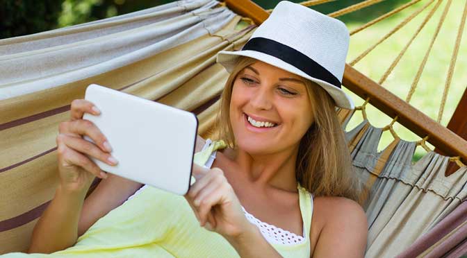 Woman in a white hat and yellow dress lies in a hammock, smiling while using her tablet to research 50-50 college lists.