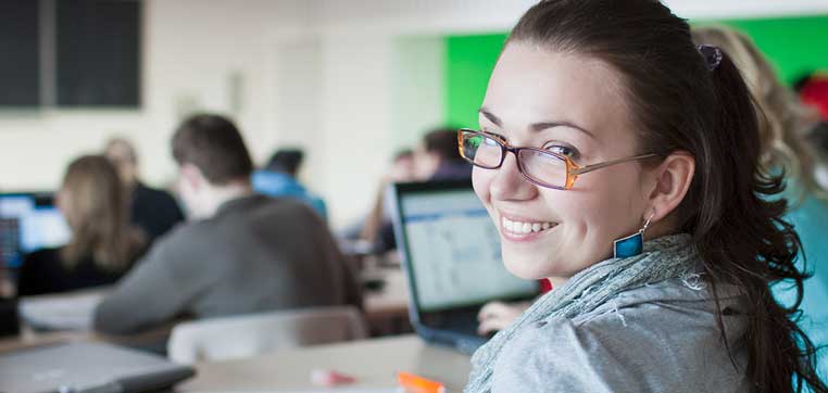 A woman in glasses smiles at the camera while sitting in a classroom with other students using laptops in the background, likely discussing their TuitionFit financial aid award letters.