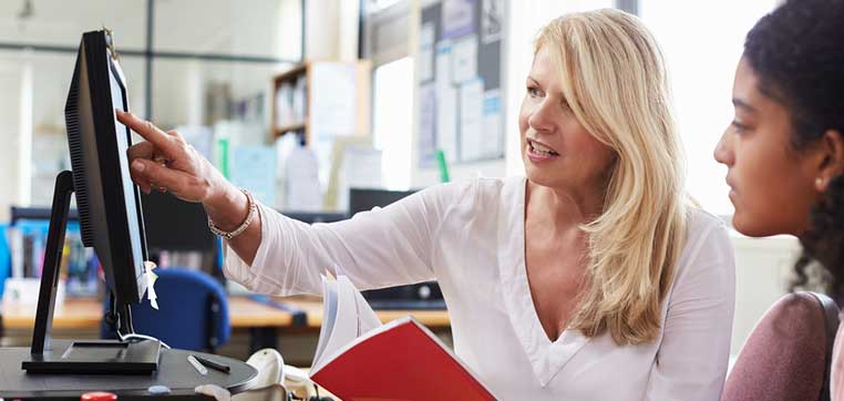 A woman with blonde hair, wearing a white blouse, points at a computer screen while holding a red notebook. Another person, with dark curly hair, looks on attentively. They are in an office setting, comparing college counseling options.