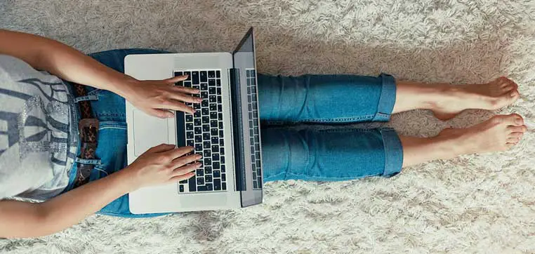 Person sitting on the floor with legs stretched out, typing on a laptop placed on their lap. They are wearing jeans and a t-shirt, contemplating the pros and cons of their Big Future. The surface appears to be a light-colored carpet.