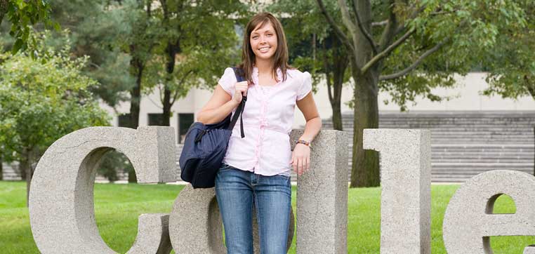 A woman stands outdoors, smiling and leaning against a large stone sculpture with the text "Calle." She is holding a backpack and is dressed casually. Trees and a building are visible in the background, making it an ideal spot for some summer college prep tips.