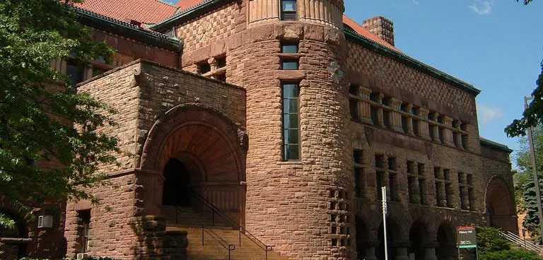 A large, historic red-brick building with arched entrances and a tower, surrounded by trees, under a clear sky, reminiscent of the classic architecture found at the University of Minnesota.