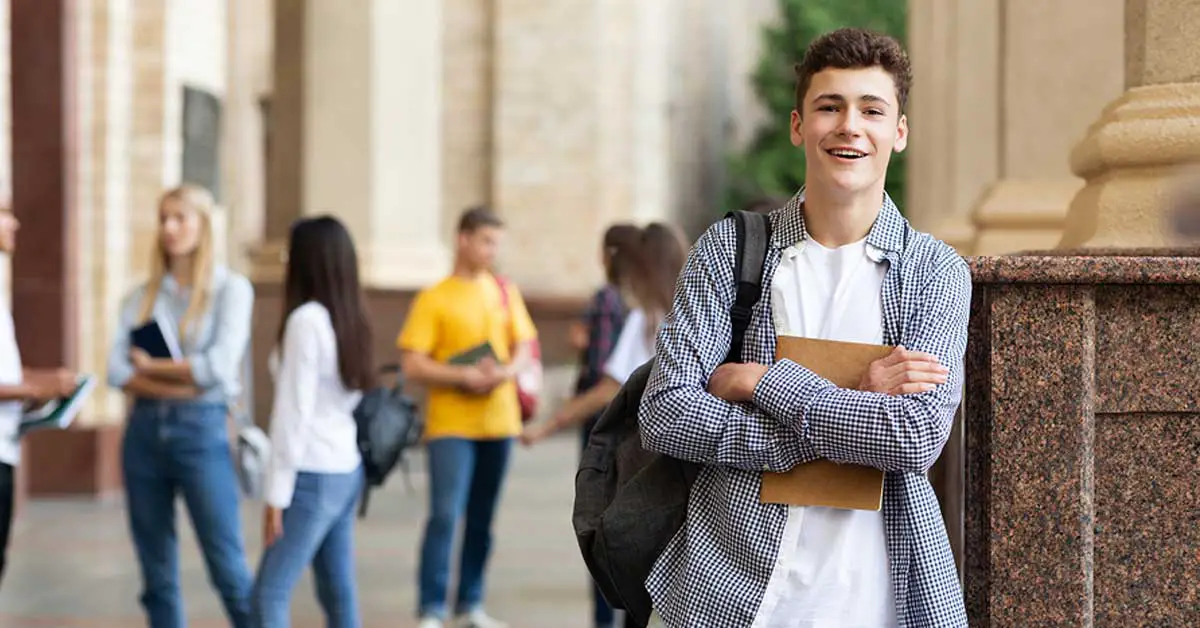 A young man stands smiling with arms crossed holding a notebook, perhaps his latest notes on researching a college. He is wearing a checked shirt over a white t-shirt with a backpack. Other students are visible in the background.